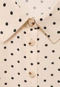 Chemise beige clair avec un motif à pois noirs, un col, et une patte de boutonnage avec un bouton rond et translucide.