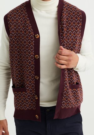 Mann trägt eine burgunderrote, gemusterte, geknöpfte Weste über einem weißen Rollkragenpullover, hält den Saum der Weste, blaue Hose sichtbar.