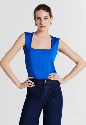 Top blu senza maniche con scollo quadrato, realizzato in tessuto liscio. Abbinato a jeans in denim scuri a vita alta con tasche posteriori.
