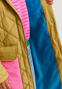 Prešívaný dlhý kabát v horčicovožltej farbe s ružovou kapsou. Interiér podšitý modrou látkou. Pod ním je oblečená ružovo pruhovaná košeľa.