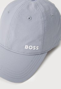 Sivá bejzbolska kapa iz lahkega materiala, ki ima beli logotip "BOSS", šivane panele in prezračevalne luknje. Oblika z zaobljenim robom.