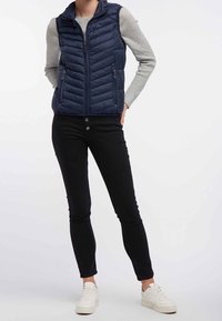 Marineblauw donsvest met een ritssluiting aan de voorkant, horizontale doorgestikte textuur en twee voorzakken. Draagt over een lichtgrijze trui en zwarte broek.