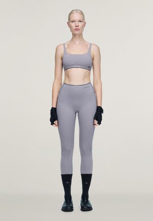 Femme debout, face à l’avant, portant un soutien-gorge de sport gris et un legging, des gants noirs sans doigts, des chaussettes noires et des chaussures de sport noires sur un fond clair.
