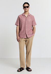 Homme portant une chemise rose à manches courtes à motifs, un pantalon beige texturé, des sandales tressées noires et des lunettes de soleil noires, se tenant devant un fond uni.
