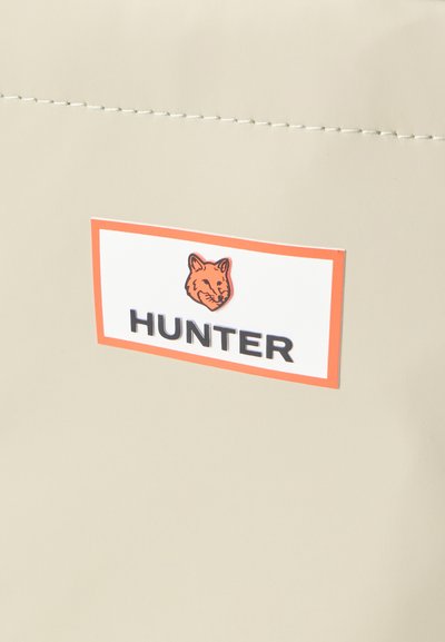 Produto bege com uma textura suave, apresentando um rótulo em destaque. O rótulo tem uma borda laranja, uma ilustração de uma raposa e texto preto "HUNTER."