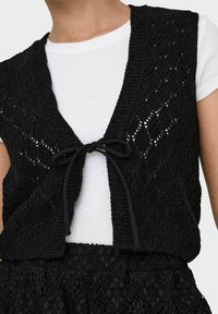 Must mustriga vest, millel on teemantmuster, avatud esiosa ja lipsusulgur, kombineeritud valge T-särgiga. Tekstuuriga kangas ja kihiline disain.
