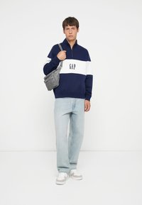 Tmavo modrý fleece pulóver s bielym prúžkom a logom "GAP"; svetlomodré džínsy; šedá crossbody taška; biele tenisky so šedými akcentmi.