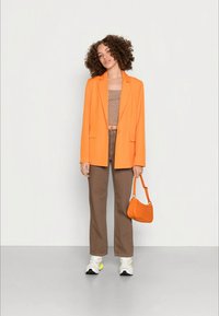 Mulher com cabelo encaracolado vestindo um blazer laranja, uma blusa curta padrão, calças castanhas e ténis brancos, segurando uma bolsa laranja contra um fundo simples.