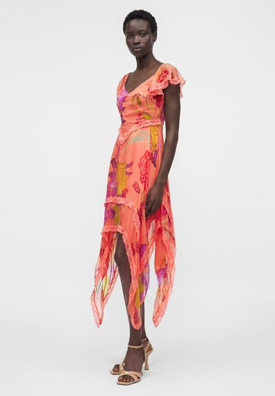 Γυναίκα με αμάνικο ασύμμετρο πορτοκαλί φόρεμα με λουλουδάτα σχέδια και δαντελένια άκρα και μπεζ ψηλοτάκουνα σανδάλια, στέκεται μπροστά σε απλό φόντο.