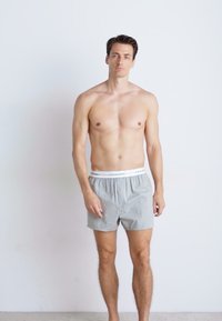 Graue Baumwoll-Boxershorts mit einem elastischen Bund, der mit "Calvin Klein" beschriftet ist. Das Modell steht aufrecht mit neutralem Gesichtsausdruck vor einem schlichten Hintergrund.