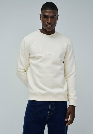Un jeune homme portant un sweat-shirt crème avec "SALSA JEANS" embossé et un jean bleu foncé, se tenant contre un fond gris clair uni.