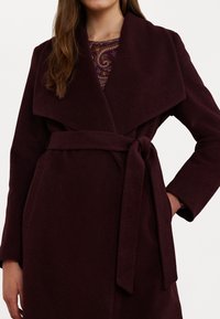 Cappotto bordeaux con ampio colletto a scialle, vita con cintura e tasche laterali. Tessuto liscio e strutturato; maniche lunghe. Strato interno decorato visibile.