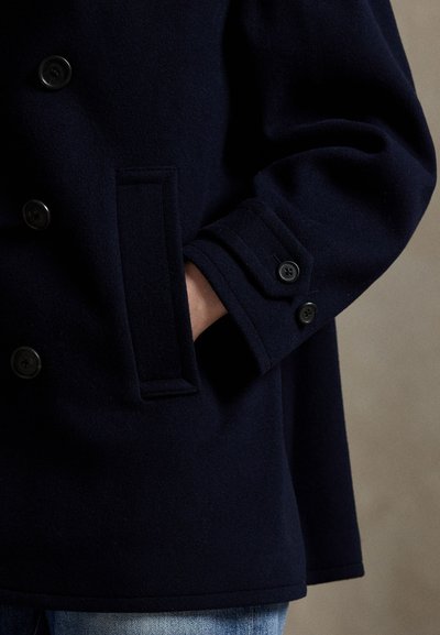 Isik kannab tumedat mereväe manteliga, millel on nupud ja küljetasku, käsi osaliselt taskus, kokku sobitatud siniste teksapükstega neutraalsel taustal.