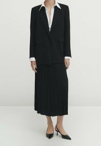 Zwarte maatwerk blazer met pieklapels, gecombineerd met een witte blouse met kraag en een geplooide zwarte midi rok. Zwarte pointy toe hakken maken de outfit compleet.