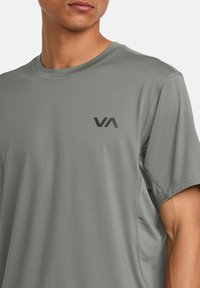 Camisa atlética de manga corta en tela gris claro con cuello redondo, que presenta un logo negro en el pecho y paneles laterales texturizados.