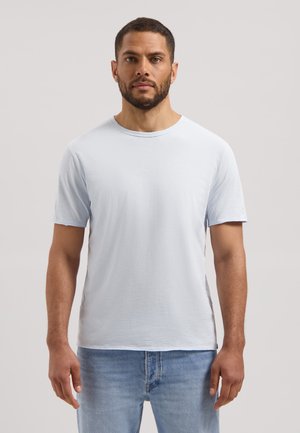 Mand med skæg iført ensfarvet lyseblå T-shirt med korte ærmer og lyseblå jeans, stående foran en ensfarvet lys baggrund.