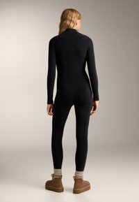 Čierny overal s dlhým rukávom, priliehavým strihom a vysokým golierom, vyrobený z elastickej tkaniny. Štýlovo doplnený hnedými čižmami a pletenými manžetami.