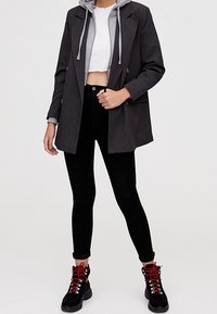 Blazer croisé gris foncé porté sur un sweat à capuche gris, associé à un jean skinny noir et des bottes noires avec des lacets rouges. Design minimaliste.
