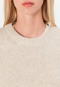 Pull en maille beige avec un col rond à côtes, présentant un motif texturé et une couche intérieure lisse, conçu pour le confort.