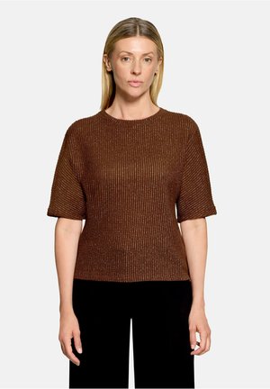 Kvinde med glat blondt hår iført en brun tekstureret kortærmet sweater og sorte bukser, stående mod en ensartet lys baggrund.