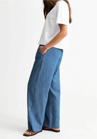 Bílé tričko s krátkým rukávem v kombinaci s volnými modrými džínovými kalhotami, které mají boční kapsy, a doplněno o ploché sandály.