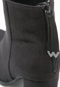 Botín de ante negro hasta el tobillo con un tacón grueso, cremallera lateral y un acabado texturizado. Presenta un diseño elegante y acentos de hardware mínimos.