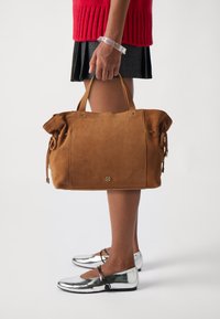 Sac fourre-tout en daim marron avec des poignées roulées, présentant des accessoires en or, des cordons latéraux et une base plate pour la stabilité. Des ballerines argentées en dessous.