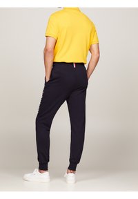 Marineblaue Sweatpants mit elastischen Bündchen, Seitentaschen und weißem Branding am linken Bein, kombiniert mit einem gelben Poloshirt und weißen Turnschuhen.
