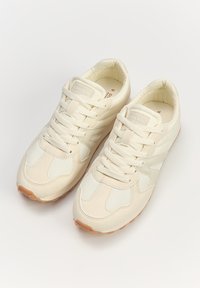 Beżowe sneakersy z materiałową i siateczkową cholewką, z teksturowanym wzorem oraz gumową podeszwą. Sznurówki i wyściełany kołnierz dopełniają projekt.