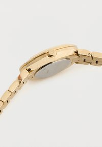 Ρολόι χειρός με χρυσό τόνο, με συνδεδεμένο βραχιόλι, πλαϊνό κορώνα με χάραξη "ck" και γυαλιστερή ασημένια πίσω πλάκα πάνω σε λευκό φόντο.