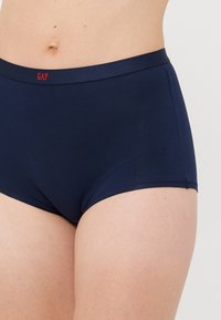 Tumesinise puuvillased aluspüksid punase "GAP" logoga vööl, kantud heleda nahaga inimesel.