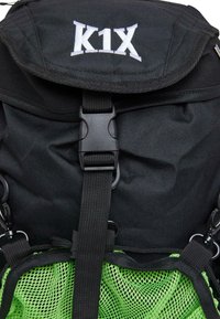Svart ryggsäck med vit "K1X" logotyp, som har ett topplock, svarta spännen och en grön meshficka i botten för extra förvaring.