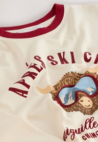 Kremna majica s kapuco z bordo rdečim okroglim izrezom in grafičnim potiskom ovce, ki nosi modna sončna očala, z napisom "APRÈS SKI" v močnih rdečih črkah.