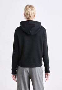 Svart hettegensere med en avslappet passform, med et enkelt design, ribbede mansjetter og nedre kant. Båret med grå bukser.