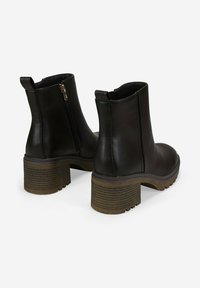 Par de botas negras hasta el tobillo con tacones de bloque apilados, cremallera lateral en una bota y tiradores en la parte trasera sobre fondo blanco.