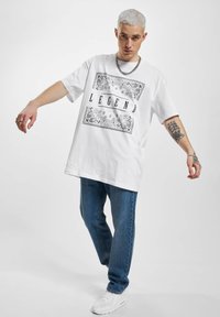 Hvit t-skjorte med svart "LEGEND" grafikk og intrikat kantdesign, sammen med blå denimjeans og hvite joggesko.
