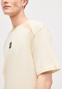 Άνδρας που φοράει μπεζ κοντομάνικη μπλούζα με μικρό μαύρο λογότυπο στην περιοχή του στήθους και ταμπά πατς στον ώμο και το τελείωμα του μανικιού.