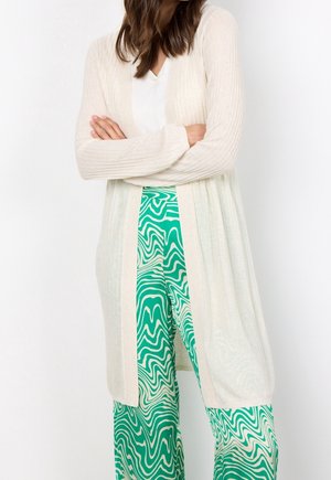 Frau trägt eine lange beige Strickjacke über einem weißen Top und grünen, wellenförmig gemusterten, weiten Hosen, Arme vor der Taille verschränkt.