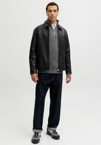 Vīrietis stāv, ģērbies melnā ādas jakā, pelēkā rāvējslēdzēja džemperī, tumšās džinsās un pelēkās sporta kurpēs uz vienkārša fona.