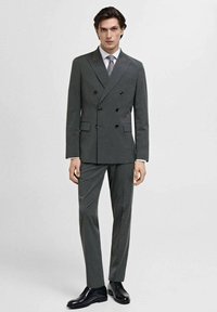 Abito grigio doppiopetto con rever a punta, due tasche frontali e bottoni neri. Abbinato a una camicia bianca e una cravatta grigia. Scarpe nere lucide.