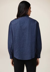 Camicia in denim con maniche lunghe e gonfiate, orlo arrotondato e un motivo discreto di piccoli punti metallici su uno sfondo blu scuro.