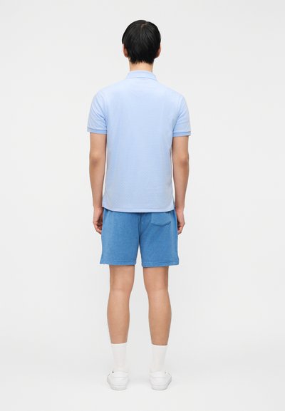 Άνδρας που στέκεται γυρισμένος με την πλάτη φοράει γαλάζιο πόλο με υφή, μπλε σορτς με πίσω τσέπη, λευκές κάλτσες και λευκά αθλητικά παπούτσια.