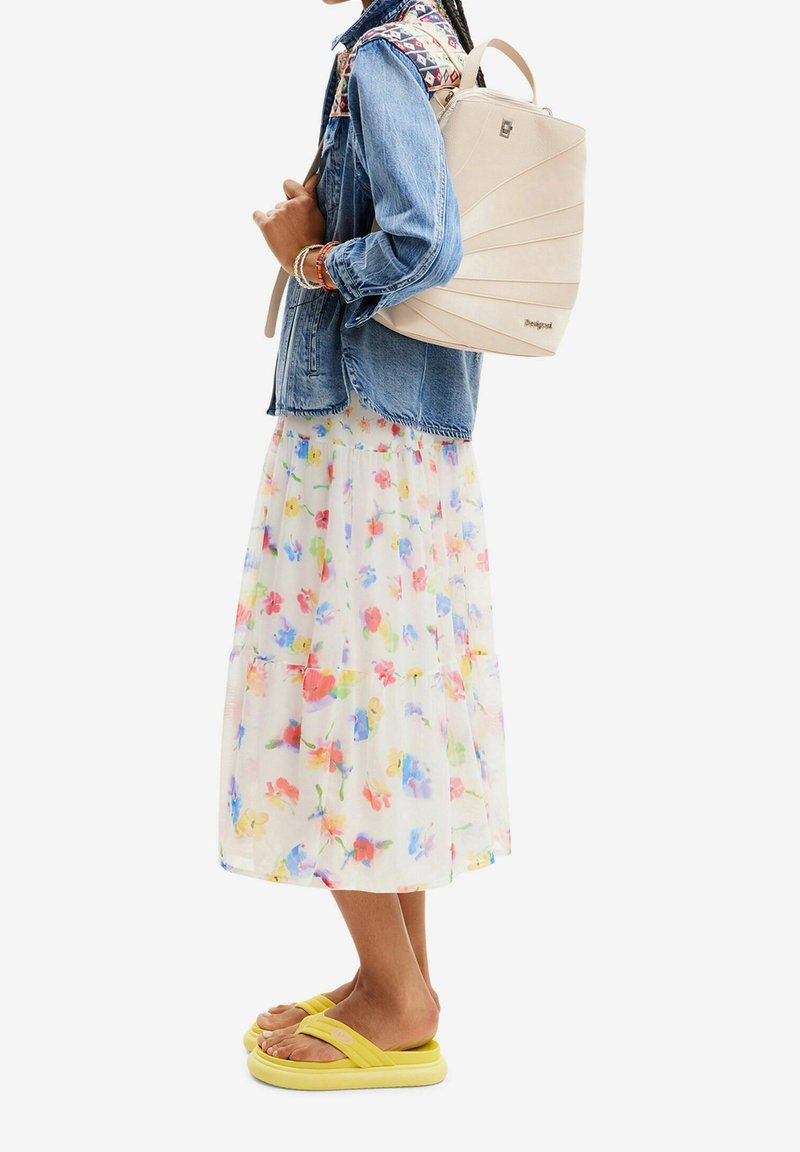 Femme portant une jupe fleurie, une veste en jean avec un empiècement à motif, des sandales jaunes et portant un sac à dos beige dans le dos.