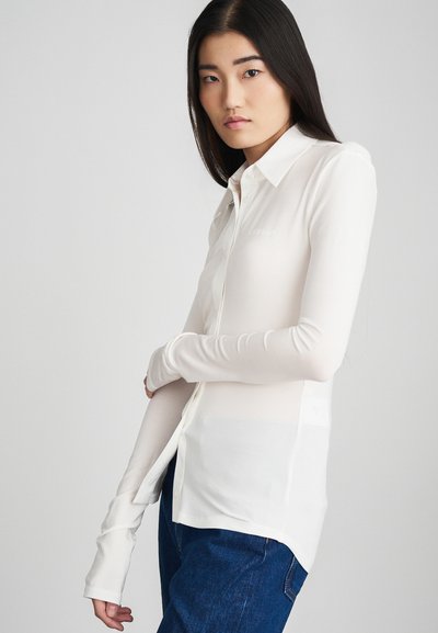 Camicia bianca a maniche lunghe con bottoni, realizzata in un tessuto morbido ed elastico, dotata di colletto e di un logo discreto sul petto, abbinata a jeans blu.