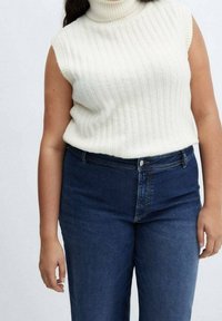 Roomkleurige geribbelde mouwloze coltrui gecombineerd met donkerblauwe jeans met hoge taille. Zachte textuur met een nauwsluitende pasvorm bij de hals.