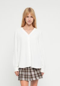 Chemise blanche  à manches longues avec boutons, col en V, tissu texturé et coupe décontractée, assortie à une jupe plissée à carreaux dans des tons marron et gris.