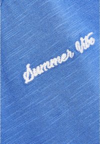 Niebieska bawełniana tkanina z teksturowanym wykończeniem, z białym haftowanym napisem "Lato '96" umieszczonym diagonalnie na powierzchni.