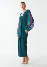 Tarnfarbendes, langärmliges Kleid mit V-Ausschnitt und strukturiertem Stoff. Verfügt über transparente, Ballonärmel und eine taillierte Detail mit Schleife. Getragen mit weißen Riemchen-Heels.