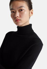 Schwarzer Rollkragenpullover mit geripptem Ausschnitt und Struktur, die auf einen weichen Stoff hinweist. Nahaufnahme des Gesichts des Modells vor neutralem Hintergrund.