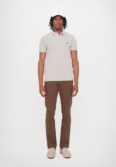 Polo gri cu logo verde, pantaloni maro și mocasini bej. O șapcă albă completează ținuta. Design simplu, casual.